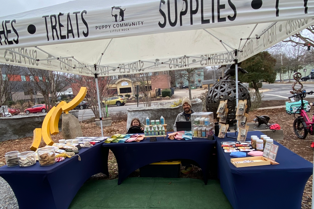 a dad and daughter sit under a market tent surrounded by puppy community brand dog treats. there are three tables loaded up with goodies with a turf carpet between.