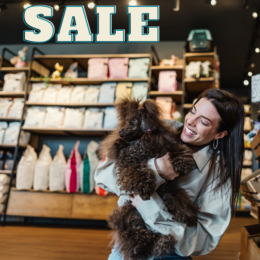 a woman smiles at her little dog standing in front of a sale section in a pet store