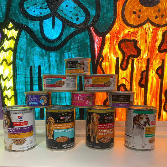 Assorted cans of pet food on a table with a colorful abstract painting of the Puppy Community window in the background.